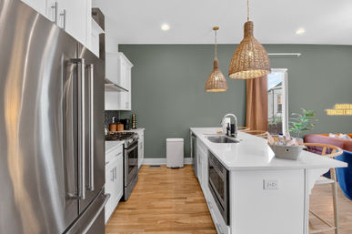 Open-concept kitchen with white island and sink, stainless steel fridge, rattan pendant lights, sage green accent wall and hardwood floors.