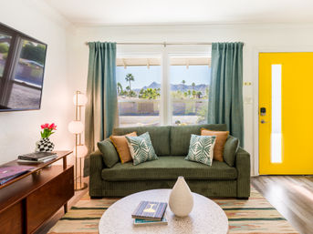 Bright mid-century living room with green corduroy sofa and patterned pillows, yellow front door, round coffee table with vase and books, and a large window framing palm trees and distant mountains.