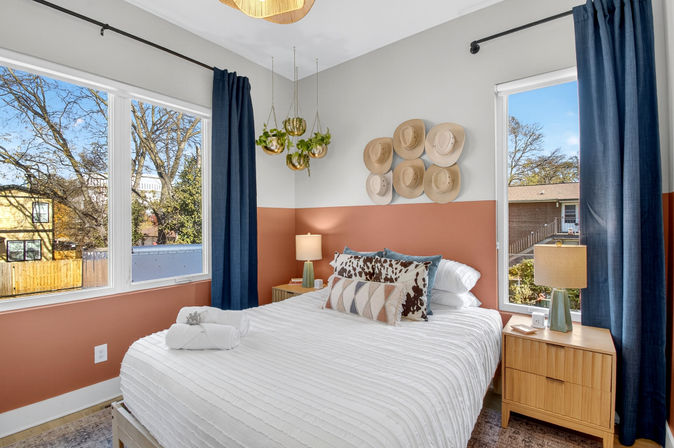 Bright modern bedroom with large windows, terracotta half-wall paint, navy curtains, white textured bedding with patterned and cowhide pillows, wood nightstands with lamps, hanging planters and a decorative wall of straw hats.