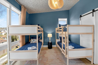 Cozy coastal-style bedroom with two natural wood twin bunk beds sleeping four, navy bedding, large windows with beige curtains, woven pendant light and bedside lamp against deep blue walls.