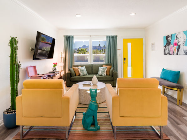 Bright mid-century modern living room with a yellow door, green sofa, two mustard armchairs, teal geometric dog statue, tall cactus, wall-mounted TV, record player, and a window framing palm trees and mountains.