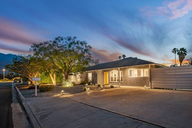 Single-story desert home at dusk with warm entry light, illuminated xeriscape landscaping, wide concrete driveway, mailbox, palm trees and mountain silhouette beneath a colorful twilight sky.