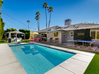 Sunny Palm Springs backyard with rectangular swimming pool, poolside cabana, pink dining set under a yellow umbrella, ping-pong table and tall palm trees.