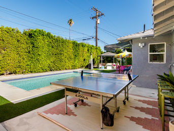 Sunlit poolside suburban backyard with a ping-pong table on a patio rug, colorful umbrella and lounge seating beside tall privacy hedges and a lone palm tree.