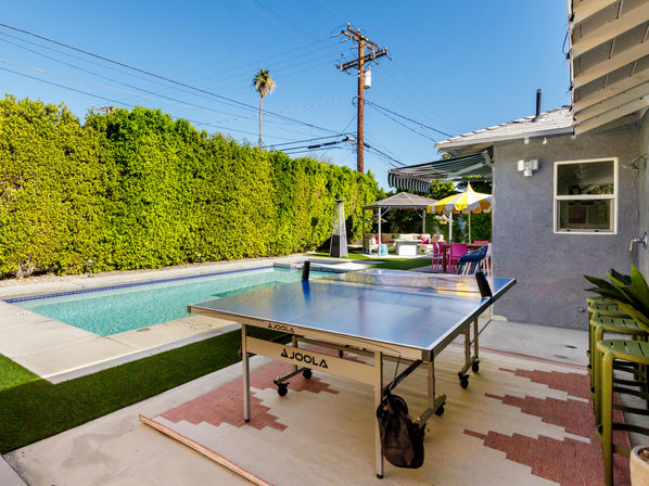 Sunlit poolside suburban backyard with a ping-pong table on a patio rug, colorful umbrella and lounge seating beside tall privacy hedges and a lone palm tree.