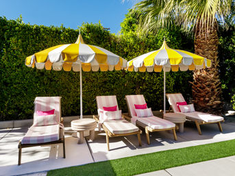 Sunny backyard poolside scene with four pink-and-white striped lounge chairs, bright yellow scalloped umbrellas, round side tables, a palm tree and a manicured green hedge.