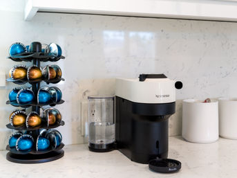 Sleek black-and-white single-serve coffee machine with clear water tank on a white marble kitchen counter, flanked by a rotating rack of metallic blue and gold coffee capsules and two white ceramic canisters in a modern home kitchen.