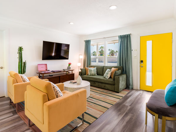 Sunny mid-century-modern living room with a bright yellow front door, green sofa, mustard armchairs, wall-mounted TV, pink turntable, cactus, and palm trees visible through the window.