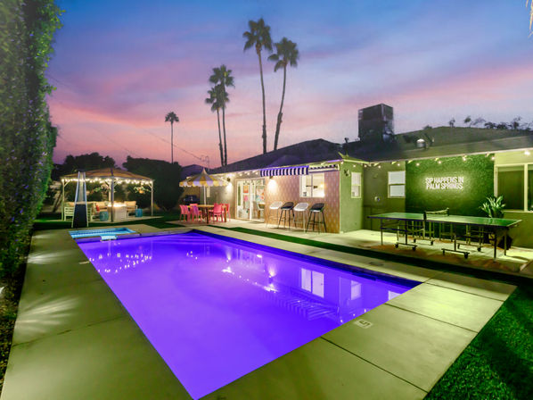 Palm Springs backyard at twilight with a purple-lit rectangular pool and spa, cozy gazebo lounge, colorful patio dining under an umbrella, bar seating, ping-pong table, string lights and tall palm trees