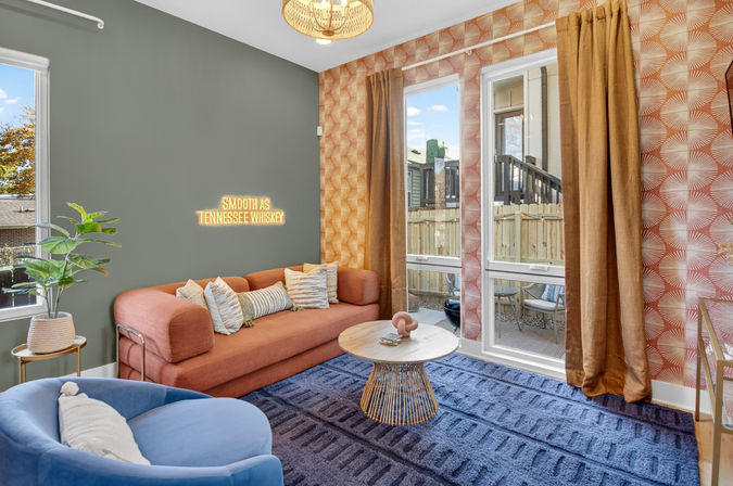 Cozy modern living room with a coral sofa, blue round accent chair, navy textured rug, rattan coffee table, potted plant, orange geometric wallpaper, tall windows to a patio, and a neon sign that reads 'Smooth as Tennessee Whiskey'.