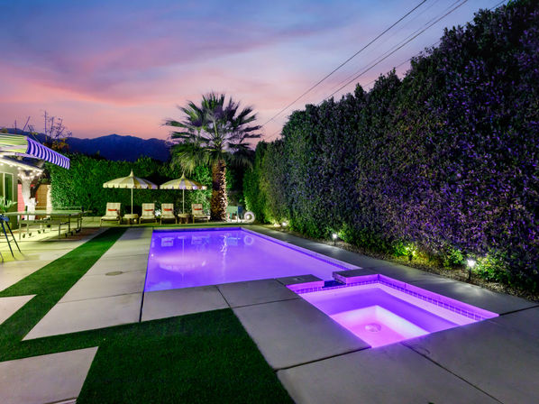Twilight backyard pool and attached spa glowing purple, palm tree with lounge chairs and striped umbrellas, tall privacy hedges and distant mountain silhouette under a pink‑purple sky.