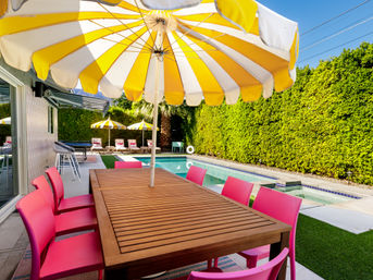 Sunny backyard pool patio with a large yellow-and-white umbrella over a wooden dining table surrounded by bright pink chairs, poolside loungers with matching umbrellas, a palm tree and tall green hedges.