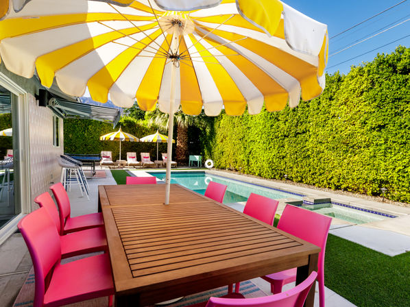Sunny backyard pool patio with a large yellow-and-white umbrella over a wooden dining table surrounded by bright pink chairs, poolside loungers with matching umbrellas, a palm tree and tall green hedges.