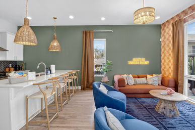 Cozy open-plan living room and kitchen with green accent wall, rust-orange sofa, blue velvet chairs, rattan pendant lights, wooden breakfast-bar stools, and neon 'Smooth as Tennessee Whiskey' sign.