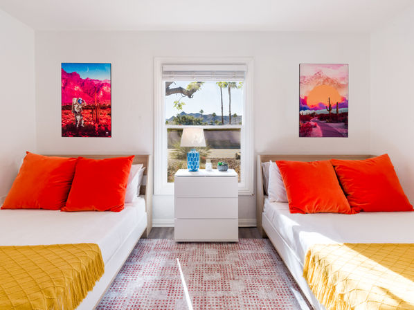 Vibrant twin bedroom with two white beds, bold orange pillows and mustard throws, white nightstand with blue lamp, window framing palm trees and desert mountains, colorful desert-themed wall art.