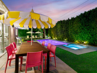 Vibrant residential backyard patio at twilight with a wooden dining table under a yellow-and-white striped umbrella, six pink chairs, illuminated swimming pool and spa, lounge chairs and tall hedge privacy wall.