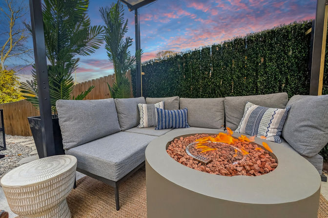 Cozy backyard patio with gray sectional sofa, patterned throw pillows and a round fire pit filled with lava rocks, framed by potted palms, a privacy hedge and pink sunset sky.