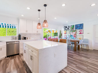 Bright modern open-plan kitchen with a large white quartz island, woven pendant lights, stainless dishwasher, wood-look floors, and a colorful dining area opening to a sunny patio.