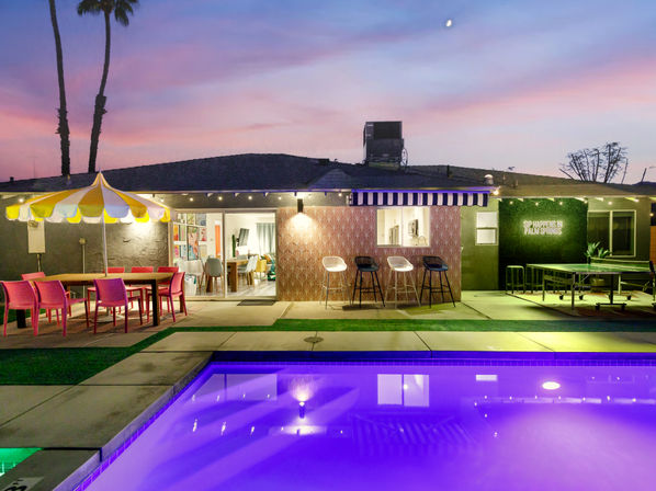 Vibrant Palm Springs backyard at sunset with a glowing purple pool, colorful patio umbrella and dining set, bar counter with stools under a striped awning, string lights and silhouetted palm trees.