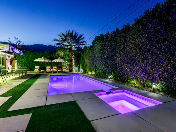 Nighttime backyard pool and spa glowing purple with LED lights, concrete decking and turf strips, lounge chairs with striped umbrellas, palm tree and tall privacy hedge against a mountain silhouette at dusk.