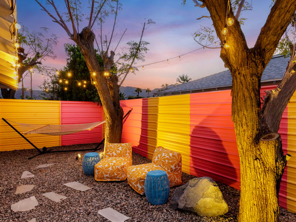 Colorful backyard patio at twilight with string lights, hammock, orange floral lounge chairs, blue wicker stools, pink and yellow corrugated fence, gravel stepping-stone path and silhouetted mountains.