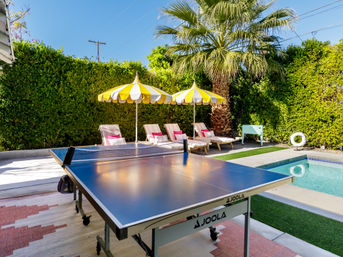 Bright sunny backyard with a table-tennis table by a turquoise pool, palm tree, yellow-and-white striped umbrellas and cushioned lounge chairs.