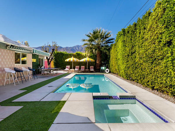 Resort-style backyard with rectangular swimming pool and built-in hot tub, palm tree, yellow-striped umbrellas and mountain backdrop under a clear blue sky