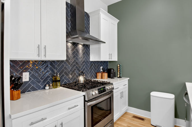 Contemporary home kitchen with white shaker cabinets, stainless steel gas range and hood, glossy blue herringbone tile backsplash, quartz countertops, wooden knife block, kettle, coffee station and green accent wall.