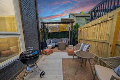 Cozy backyard patio with pergola and string lights, gray sectional around a fire pit, wicker chairs, small charcoal grill, wooden privacy fence and colorful sunset sky.