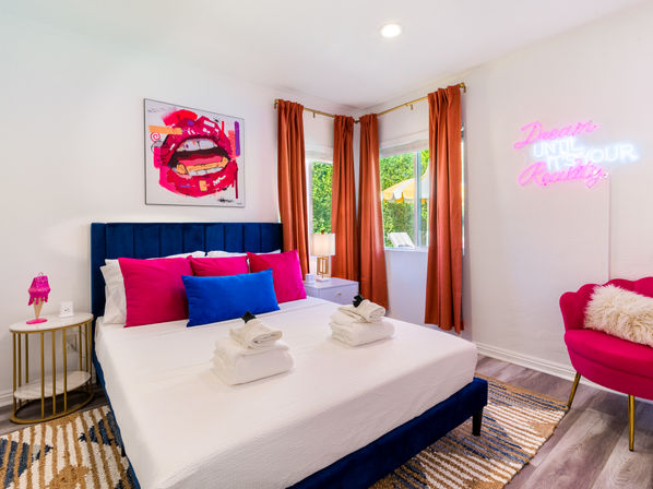 Sunlit modern bedroom with blue velvet bed, hot-pink and royal-blue pillows, pop-art lips artwork, pink neon "Dream Until It's Your Reality" sign, and rust-orange curtains.