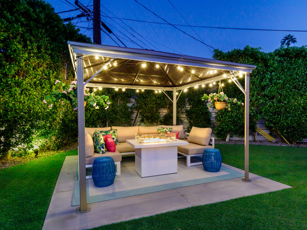 Cozy backyard gazebo at dusk with string lights, L-shaped outdoor sofa with colorful cushions, fire-pit table, hanging plant baskets and lush hedges