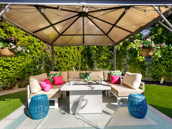 Sunlit backyard garden gazebo with a cozy beige sectional and bright pink and tropical-print pillows around a central fire-pit table, blue wicker stools, hanging flower baskets and string lights against a green hedge.
