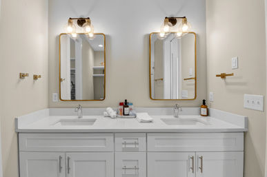 Modern bathroom double vanity with white quartz countertop, dual undermount sinks, brass-framed mirrors, globe wall sconces, folded towels and soap bottles on a neutral wall