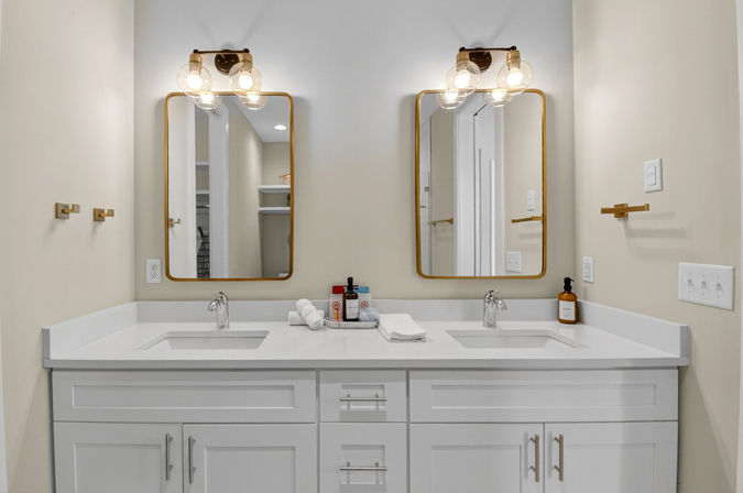 Modern bathroom double vanity with white quartz countertop, dual undermount sinks, brass-framed mirrors, globe wall sconces, folded towels and soap bottles on a neutral wall