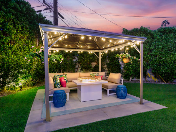 Cozy suburban backyard gazebo at sunset with twinkling string lights, cushioned L-shaped sofa, fire-pit table, hanging flower baskets and tall privacy hedges.
