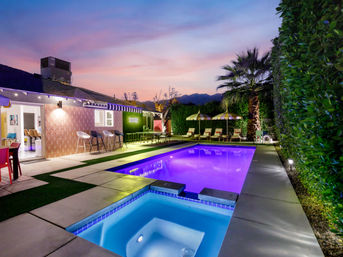 Sunset backyard patio with a glowing purple rectangular pool and blue-lit spa, striped umbrellas and lounge chairs, palm tree and mountain silhouette