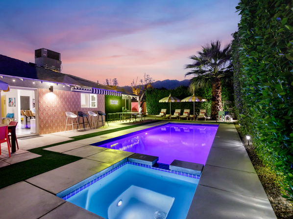Sunset backyard patio with a glowing purple rectangular pool and blue-lit spa, striped umbrellas and lounge chairs, palm tree and mountain silhouette