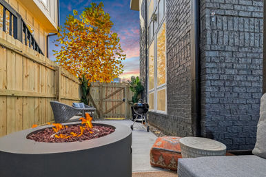 Cozy narrow backyard patio with modern round fire pit, wicker seating, charcoal grill, brick exterior, wooden fence and autumn tree under a colorful sunset sky.