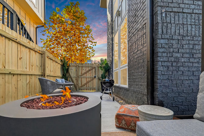 Cozy narrow backyard patio with modern round fire pit, wicker seating, charcoal grill, brick exterior, wooden fence and autumn tree under a colorful sunset sky.