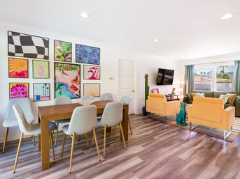Sunny modern open-plan living and dining room with wood floors, vibrant gallery wall, gray dining chairs, mustard-yellow armchairs and large windows showing palm trees.