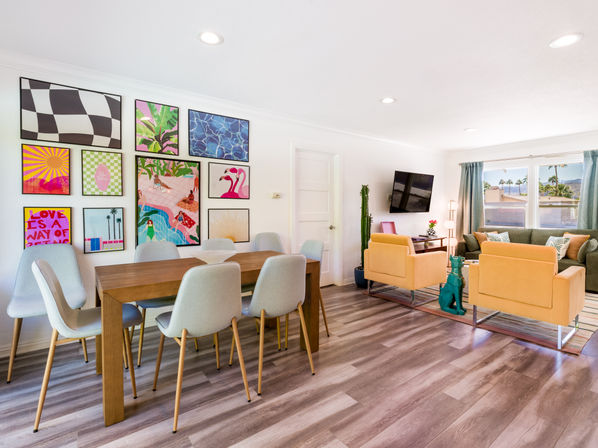 Sunny modern open-plan living and dining room with wood floors, vibrant gallery wall, gray dining chairs, mustard-yellow armchairs and large windows showing palm trees.