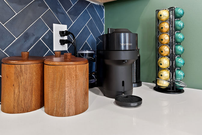 Kitchen countertop coffee station with a black espresso machine, vertical rack of gold and teal coffee pods, two wooden canisters, white countertop and navy herringbone tile backsplash