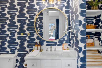 Modern coastal-style bathroom with navy brushstroke wallpaper, round gold-framed mirror over a white vanity with chrome faucet, toiletries on the countertop and built-in open shelves holding towels and decor.