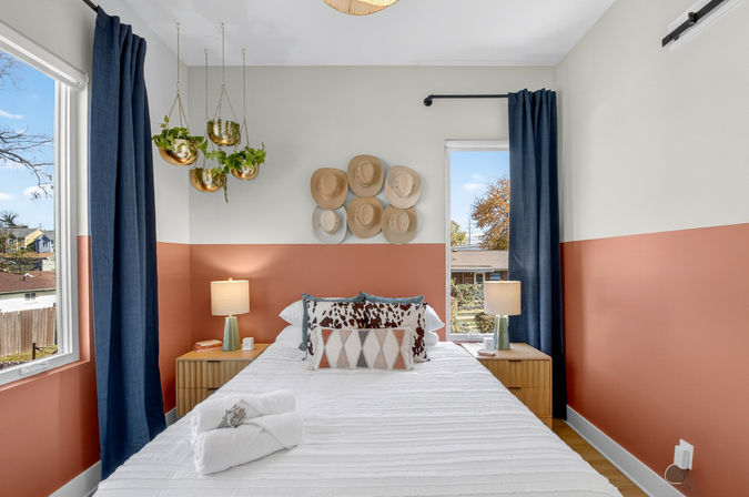 Cozy modern bedroom in a sunny suburban home with a terracotta half-wall, white bedding, navy curtains, hanging gold planters, a playful wall of straw hats, and wooden nightstands with lamps.
