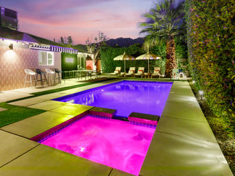 Colorful backyard pool and hot tub glowing purple and pink at dusk, modern patio with lounge chairs, umbrellas, bar stools, palm tree and mountain silhouette.