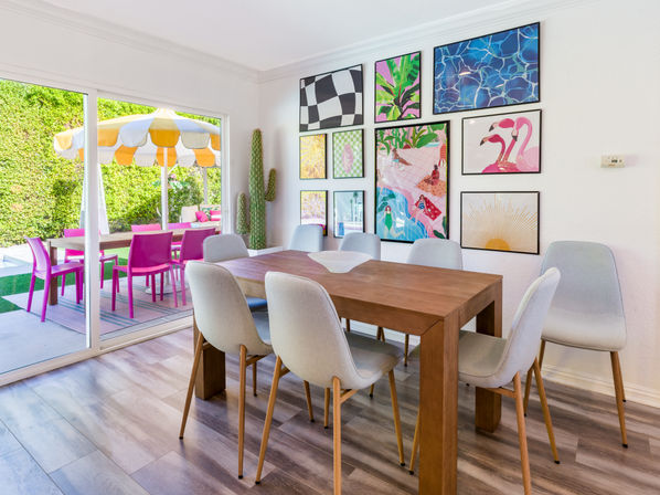 Sunlit modern dining room with wooden table and six light-gray chairs, colorful gallery wall and tall cactus, sliding glass doors opening to a sunny backyard patio with a yellow-and-white umbrella and bright pink outdoor chairs.