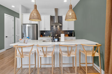 Open-concept modern kitchen with white island, four light-wood bar stools, woven rattan pendant lights, stainless steel refrigerator and herringbone navy tile backsplash on a sage-green wall.