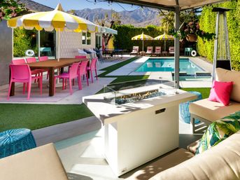 Sunny backyard pool patio with yellow-and-white umbrellas, bright pink dining chairs, cabana fire table, lounge seating, ping-pong table and desert mountain backdrop.