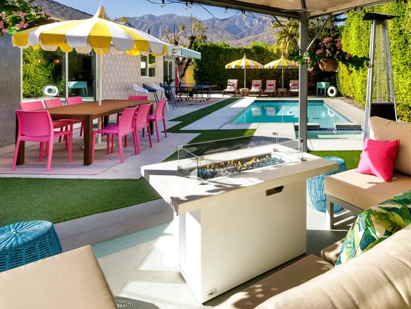 Sunny backyard pool patio with yellow-and-white umbrellas, bright pink dining chairs, cabana fire table, lounge seating, ping-pong table and desert mountain backdrop.