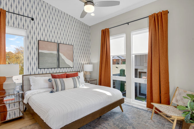 Sunlit modern bedroom with geometric patterned accent wall, white textured bedding and decorative pillows, rust-orange curtains framing tall windows, ceiling fan, bedside lamps, woven chair and leafy plant with neighboring homes visible outside.